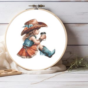 May include: Cross-stitch embroidery of a young person in a cowboy hat, holding a coffee cup. The design features a brown hat, blue top, orange skirt, and blue boots. The embroidery is set in a wooden hoop.