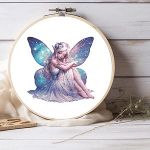 May include: A cross-stitch embroidery project in a circular wooden hoop featuring a seated fairy with long light hair and pointed ears. The fairy wears a white dress and has large, translucent wings patterned with blue, purple, and white galaxy stars.