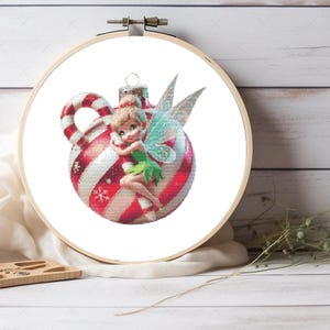 May include: A circular wooden embroidery hoop containing a cross-stitch pattern of a small fairy with blonde hair and green clothing, hugging a large, round, red and white striped Christmas ornament decorated with white snowflake accents.