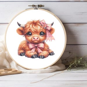 May include: A circular wooden embroidery hoop containing a cross-stitch pattern of a fluffy brown highland calf. The calf has large dark eyes, small horns, and is adorned with a light pink bow on its head and a matching pink bow around its neck.