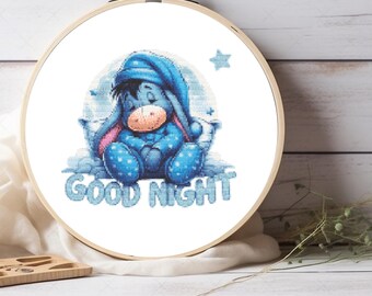 Puede incluir: Un bastidor de bordado circular de madera que contiene un patrón de punto de cruz de un burro azul con gorro de dormir y pijama a juego, sentado sobre una almohada. El texto GOOD NIGHT está bordado en letras azul claro bajo la figura.