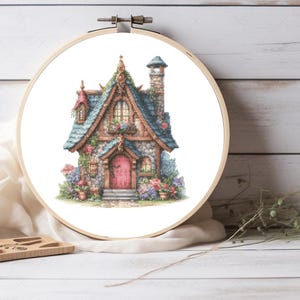 Peut inclure: Un motif de point de croix représentant un charmant chalet en pierre avec une porte rouge, un toit bleu et une cheminée. Le chalet est entouré de fleurs et de plantes.