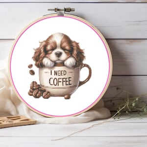 May include: Cross-stitch embroidery in a wooden hoop features a Cavalier King Charles Spaniel puppy nestled in a coffee cup. The cup reads "I NEED COFFEE" and is surrounded by coffee beans. The puppy has brown and white fur.
