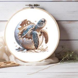 May include: A circular wooden embroidery hoop containing a cross-stitch pattern of a sea turtle hatching from a cracked egg. The turtle features shades of blue, green, and brown, while the egg is textured with white and tan tones on a white fabric.
