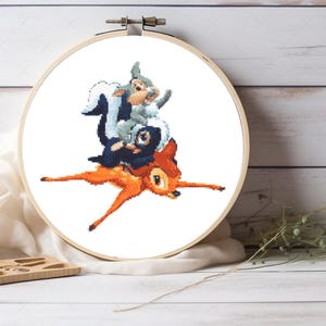 May include: A circular wooden embroidery hoop containing a cross-stitch pattern of a light brown deer with a grey rabbit and a black skunk sitting on its back. The design is centered on a white fabric background, showcasing detailed pixelated art.