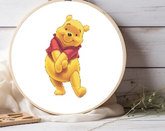 May include: A cross-stitch embroidery hoop featuring a yellow Winnie the Pooh character wearing a red shirt. The image is set against a white background with a wooden hoop and a wooden surface.