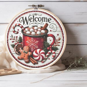 Op de afbeelding: Een ronde borduurring met een wit achtergrond en een rode mok gevuld met warme chocolademelk en marshmallows. De mok is omringd door pepermuntstokken, kaneelstokjes, een peperkoekman en een pepermuntsnoepje. De tekst "Welcome" is in zwarte cursieve letters boven de mok geschreven.