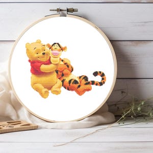 Könnte beinhalten: Ein Kreuzstich-Stickrahmen mit Winnie Puuh und Tigger. Puuh trägt ein rotes Hemd, und Tigger ist orange mit schwarzen Streifen. Die Stickerei ist vor einem weißen Hintergrund.