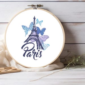 May include: A circular wooden embroidery hoop containing a cross-stitch pattern on white fabric. The design features a dark blue Eiffel Tower, the word Paris in cursive script, and several butterflies in shades of light blue and purple.