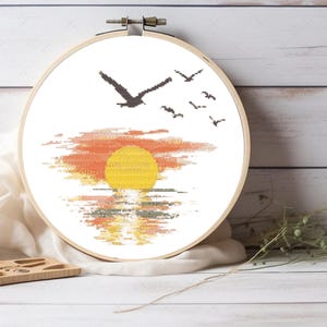 May include: A cross-stitch pattern of a sunset over the ocean with birds flying in the sky. The sun is a bright yellow and the sky is a gradient of orange, pink, and red. The ocean is a dark blue with a reflection of the sunset.