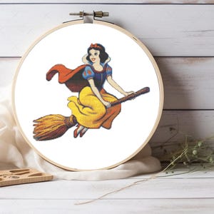 Puede incluir: Un bastidor de bordado de punto de cruz con una imagen colorida de Blancanieves montada en una escoba. El diseño incluye el vestido amarillo, la capa roja y el pelo negro de Blancanieves. El bordado está sobre un fondo blanco.
