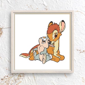 May include: A square cross-stitch pattern featuring a tan deer with brown markings and a small gray rabbit. The deer is nuzzling the rabbit, which is sitting upright. The design is rendered in a pixelated style with distinct grid lines on a white background.