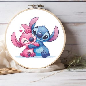 May include: A cross-stitch embroidery hoop featuring the characters Stitch and Angel from Lilo & Stitch. Stitch is blue, and Angel is pink, both embracing. The embroidery is set against a white background.