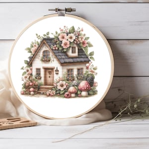 May include: A circular wooden embroidery hoop containing a fabric panel with a printed illustration of a cottage. The house features a dark roof, white walls, and a wooden door, surrounded by pink flowers, green foliage, and red spotted mushrooms.