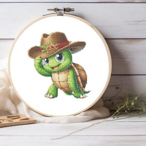 May include: Cross-stitch embroidery of a cartoon turtle wearing a brown cowboy hat with a floral accent. The turtle is green and brown, with large, friendly eyes. The embroidery is set in a wooden hoop, perfect for home decor.