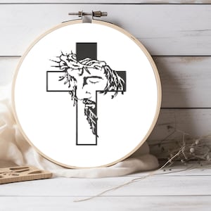 May include: A black and white cross-stitch design of Jesus Christ wearing a crown of thorns, set within a wooden embroidery hoop. The cross is centered, with the face of Jesus depicted on the upper portion. The background is white.