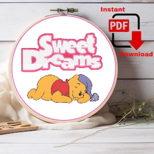 May include: A cross-stitch embroidery hoop featuring a sleeping Winnie the Pooh design. The text "Sweet Dreams" is stitched in pink, with a red PDF download icon. The design is set against a white background.