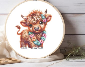 May include: A cross-stitch embroidery project in a round wooden hoop featuring a fluffy brown Highland calf with large dark eyes, small horns, and a colourful floral garland around its neck, set against a plain white fabric background.