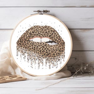 Può includere: Un cerchio da ricamo a punto croce con un disegno di labbra con un motivo leopardato. Le labbra sono leggermente aperte e il disegno cola verso il basso.