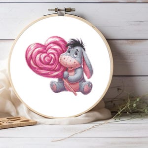 May include: A cross-stitch embroidery hoop featuring a gray donkey character holding a large pink heart-shaped lollipop. The donkey has pink accents and is set against a white background.