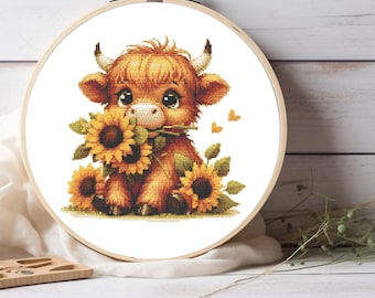 May include: A cross-stitch pattern in a round wooden embroidery hoop featuring a fluffy brown Highland calf with large dark eyes and small horns, holding a bright yellow sunflower in its mouth, surrounded by additional sunflowers and green leaves.