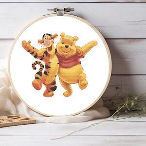 Pode incluir: Um bastidor de bordado em ponto cruz com Winnie the Pooh e Tigrão. As personagens são retratadas em amarelo, laranja e preto, com os braços estendidos. O bordado é colocado sobre um fundo branco.