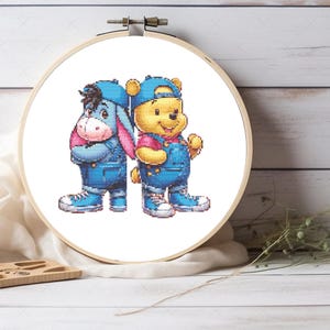 Puede incluir: Patrón de punto de cruz con Winnie de Pooh y Ígor vistiendo monos de mezclilla y gorras de béisbol. Los personajes están de pie uno al lado del otro con los brazos cruzados.