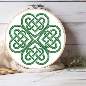 May include: A circular wooden embroidery hoop containing a cross-stitch pattern of a four-leaf clover design. The clover is composed of intricate green Celtic knotwork lines stitched onto a plain white fabric background within the frame.