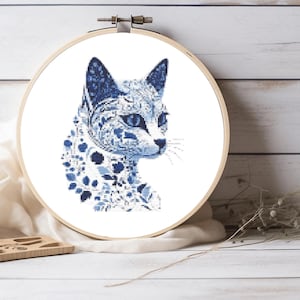 Könnte beinhalten: Ein runder Stickrahmen aus Holz mit einem Kreuzstichmuster einer Katze. Die Katze ist in Blau- und Weißtönen gehalten und weist auf ihrem gesamten Fell komplizierte Blumen- und Blattmotive auf, ergänzt durch dunkelblaue Augen und spitze Ohren.