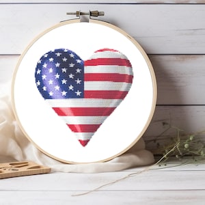 May include: A circular wooden embroidery hoop containing a cross-stitch pattern of a heart shaped like the American flag. The design features a blue field with white stars on the left and horizontal red and white stripes on the right side.