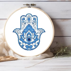 May include: A white embroidery hoop with a blue and white hamsa hand design. The hamsa hand is a symbol of protection and good luck.