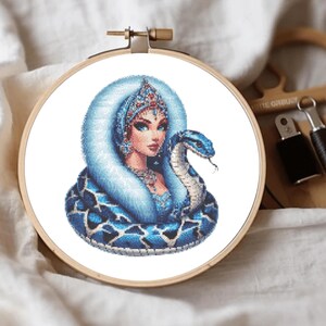 May include: A blue and white cross stitch pattern of a woman with a crown and a snake wrapped around her. The woman is wearing a blue and white dress and a white fur collar. The snake is blue and white with a black tongue.