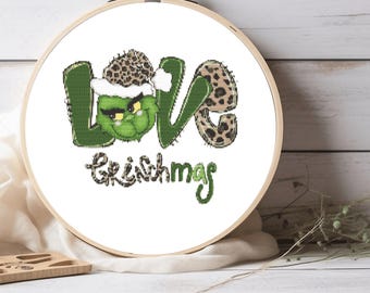Pode incluir: Um aro de bordado branco com um desenho de ponto cruz verde e marrom. O desenho diz "LOVE GRINCHMAS" com um rosto verde do Grinch usando um chapéu de Papai Noel marrom e branco no meio da palavra "LOVE".