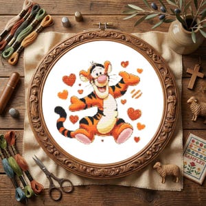 May include: A cross-stitch embroidery project featuring a cartoon tiger with orange fur and black stripes, surrounded by small orange and red hearts, displayed in a round, ornate wooden frame on a piece of white fabric.
