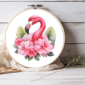 May include: A circular wooden embroidery hoop containing a cross-stitch pattern of a bright pink flamingo surrounded by large pink hibiscus flowers and green tropical palm leaves. The design is centered on a white fabric background within the frame.