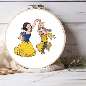 May include: A circular wooden embroidery hoop containing a cross-stitch pattern of Snow White and Dopey dancing. Snow White wears a blue bodice and yellow skirt, while Dopey wears a yellow coat and blue trousers. The design is on white fabric.