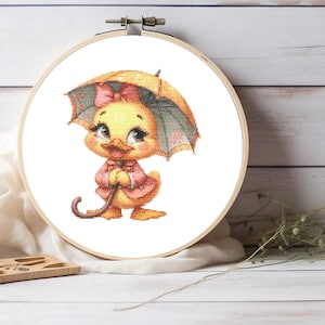 May include: A yellow duckling wearing a pink jacket holds a yellow umbrella with a pink bow. The duckling is embroidered on a white fabric.