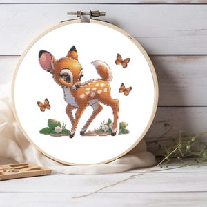 May include: A circular wooden embroidery hoop containing a cross-stitch pattern of a young deer with white spots, large dark eyes, and pink-lined ears. The deer is surrounded by three small orange butterflies and sits on a patch of green grass.