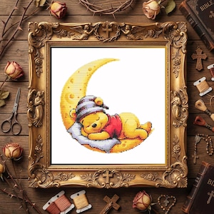 May include: A cross-stitch pattern featuring a yellow bear wearing a striped nightcap, sleeping on a crescent moon with a white pillow. The design is displayed within an ornate, rectangular gold-colored frame with intricate carvings.