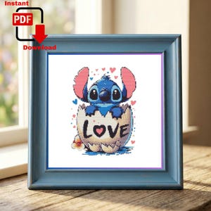 May include: A framed cross-stitch featuring a blue character emerging from an eggshell with the word "LOVE". The character has large ears and a dark blue body. The frame is a light blue colour.
