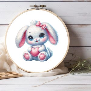 Puede incluir: Un bastidor de bordado de punto de cruz con un lindo conejito blanco sosteniendo un corazón rosa. El conejito tiene ojos azules, detalles en rosa y un lazo rosa. El bordado está sobre un fondo blanco.