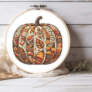 May include: A cross-stitch pumpkin design in a wooden embroidery hoop. The pumpkin features intricate patterns in shades of orange, brown, and cream, with a dark brown stem. The design includes leaves and floral elements, perfect for fall decor.