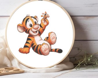 Puede incluir: Un bastidor de bordado circular de madera que contiene un patrón de punto de cruz de un cachorro de tigre de dibujos animados con rayas naranjas y negras, sentado y sosteniendo un pequeño juguete marrón. El tigre tiene vientre blanco y patas rosas.