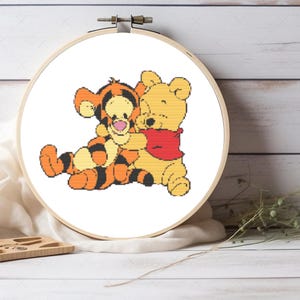 Könnte beinhalten: Ein runder Stickrahmen aus Holz mit einem Kreuzstichmuster der Figuren Tigger und Winnie Puuh. Tigger ist orange mit schwarzen Streifen und Winnie Puuh ist gelb mit einem roten Oberteil. Die beiden Figuren sind in einer Umarmung dargestellt.