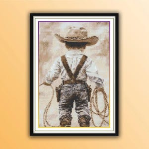 Puede incluir: Una obra de arte en punto de cruz enmarcada representa a un niño vestido de vaquero, visto por la espalda. El niño lleva un sombrero de vaquero marrón, una camisa clara, vaqueros y botas. El niño sostiene una cuerda y una funda de pistola.
