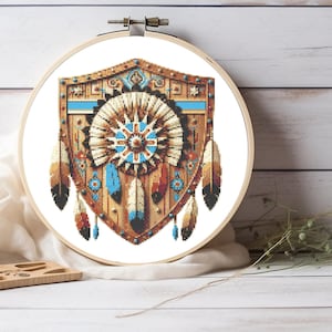 Peut inclure: Un tambour à broder circulaire en bois contenant un motif au point de croix en forme de bouclier. Le motif présente un motif central en forme de soleil, des bordures géométriques et des plumes suspendues dans des tons marron, beige, blanc et bleu vif.