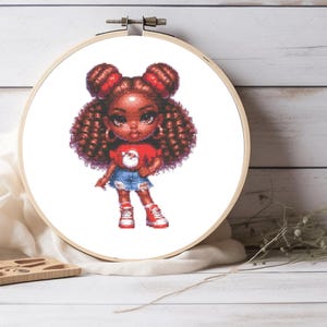May include: A cross-stitch pattern displayed in a circular wooden embroidery hoop featuring a cartoon character with dark skin, brown curly hair in two buns, wearing a red shirt with a Santa Claus graphic, blue denim shorts, and red sneakers.