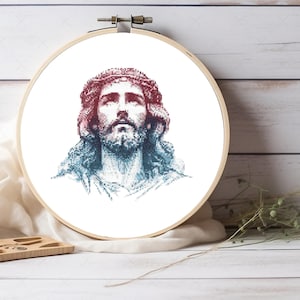 May include: A circular wooden embroidery hoop containing a cross-stitch pattern of a portrait of Jesus Christ. The image features a gradient color scheme transitioning from deep red at the top to blue at the bottom, depicting a bearded face.