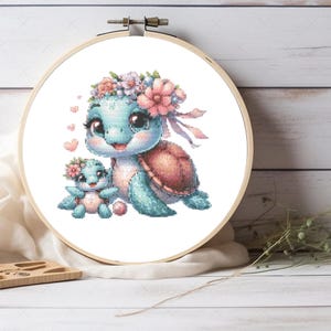 May include: A circular wooden embroidery hoop containing a cross-stitch pattern of a large light blue turtle and a smaller baby turtle. Both turtles wear pink and white floral crowns. Small pink hearts float near the larger turtle's head.