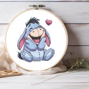 May include: A cross-stitch pattern featuring a cartoon image of Eeyore the donkey from Winnie the Pooh. Eeyore is sitting with his hands covering his face and a pink heart is floating above him.